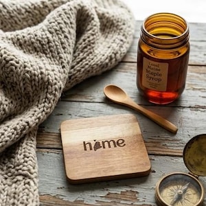 Puede incluir: Un posavasos de madera con la palabra "home" y el contorno de un estado, un tarro de jarabe de arce, una cuchara de madera y una brújula sobre una superficie de madera desgastada. Una manta de punto beige está drapeada en el fondo.