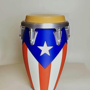 May include: A conga drum with a blue, white, and red design. The drum features the flag of Puerto Rico, with a white star on a blue background and red and white stripes. The drum has a tan drumhead and silver hardware.