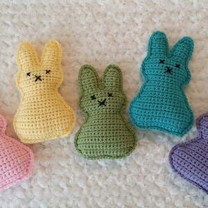 May include: Five handmade crochet bunny toys in pastel shades: pink, yellow, green, teal, and purple. Each bunny has black embroidered eyes and a stitched mouth. The bunnies are arranged on a white, textured surface.