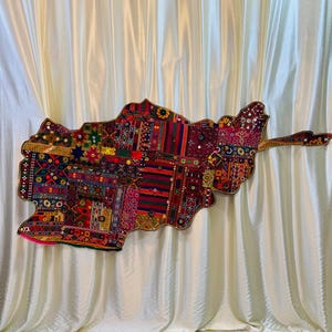 Könnte beinhalten: Ein farbenfrohes Textil-Wandbehang in Form eines Landes. Das Kunstwerk zeigt ein Patchwork-Design mit verschiedenen Mustern und Farben, darunter Rot, Blau und Gold. Die Kanten sind mit einem dekorativen Rand versehen.
