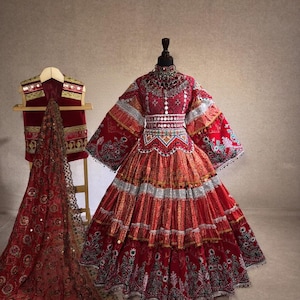 May include: A vibrant red traditional dress with intricate embroidery and embellishments. The dress features layered skirts, long sleeves, and a decorative bodice. A matching vest and shawl are also displayed, showcasing a rich cultural aesthetic.