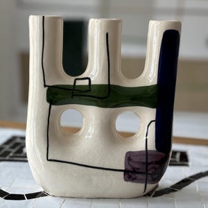 Könnte beinhalten: Weiße Keramikvase mit drei Öffnungen, verziert mit abstrakten Linien und Formen in Schwarz, Grün, Blau und Lila.