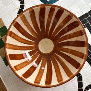 May include: A ceramic bowl with a cream-colored interior and a brown, sunburst-like pattern radiating from the center. The rim is brown, and the bowl is set against a tiled surface.