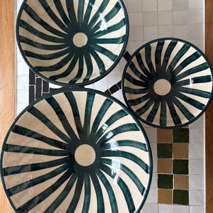 May include: Three ceramic bowls with a cream-colored interior and a dark teal spiral design. The bowls vary in size and are arranged on a mosaic tile surface. The bowls have a circular shape and a dark teal rim.