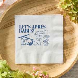 May include: White paper napkin with blue text and illustrations. The text reads "LET'S APRÈS BABES! Samantha's Bachelorette" with images of skis, a heart, and a cocktail. The napkin is on a wooden tray.