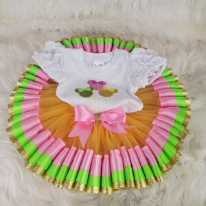 Puede incluir: Un colorido conjunto de bebé con una parte superior blanca con bordado floral y mangas de encaje. La falda tutú está en capas con tul naranja, cintas rosas y verdes y ribete dorado con purpurina. Un gran lazo rosa adorna la cintura.