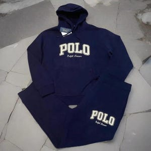 Puede incluir: Conjunto de sudadera con capucha y pantalón de chándal Polo Ralph Lauren azul marino. La sudadera tiene un bolsillo delantero y la palabra "POLO" en letras blancas. El pantalón de chándal a juego también muestra el logotipo "POLO". El conjunto es de material suave.