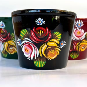 May include: Three ceramic flower pots in green, black, and burgundy, each decorated with hand-painted floral designs. The flowers have red, yellow, and white petals with green leaves. The pots are arranged on a white surface.