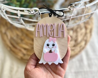 May include: Wooden oval sign with the name "AMELIA" in pink letters. A white ghost with a pink bow and pumpkin is attached to the wooden sign. The sign is hanging in front of a white basket.