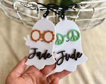 May include: Two white ghost-shaped tags with black script names "Jade" and "Jude". One ghost wears orange flower-shaped glasses, the other wears green peace sign glasses. The tags are hanging from a white basket.
