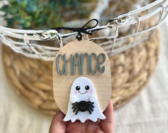 May include: An oval-shaped wooden ornament featuring the word CHANCE in raised grey lettering. Below the text is a white ghost figure with a smiling face, pink cheeks, and small arms holding a black spider. The ornament hangs from a black string.