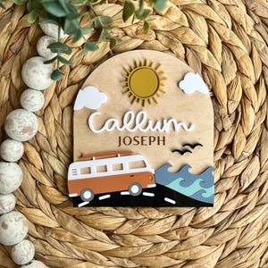 May include: Wooden sign with a beach theme. The sign features a sun, clouds, a van with a surfboard, and the name "Callum Joseph". The van is orange and white, and the waves are blue and teal.