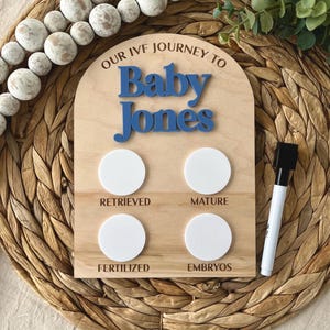 May include: Wooden IVF journey board with blue text that reads "Baby Jones". The board has four white circles and the words "Retrieved", "Mature", "Fertilized", and "Embryos". A black and white marker is included.