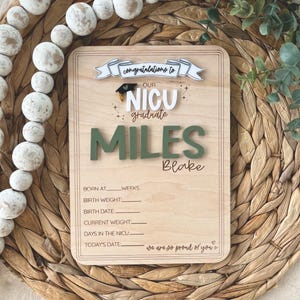 以下が含まれることがあります： 緑色の文字で「MILES」と書かれた、NICU卒業生向けの木製サイン。サインには「NICU卒業生おめでとう」というテキストと、赤ちゃんの誕生日の詳細を記入するスペースが含まれています。織り込まれた背景にあります。