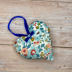 May include: A blue and white floral heart-shaped cushion with a blue velvet ribbon loop for hanging. The cushion has a light blue button with a white bow.