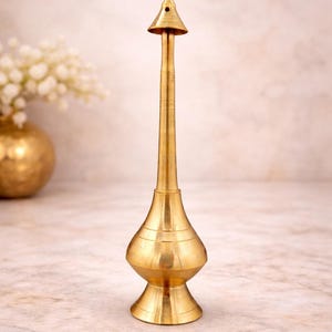 May include: A gold-coloured, ornate metal object with a conical top and a long, slender body. The base is wider, with a bulbous shape and horizontal ridges. The object is likely a decorative item or a vessel for liquids.
