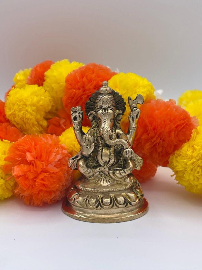 Puede incluir: Una detallada estatua de bronce de la deidad hind&uacute; Ganesha, sentado sobre una base decorativa. La estatua est&aacute; rodeada de vibrantes flores de cal&eacute;ndula naranja, amarilla y blanca. El fondo es una superficie blanca lisa.