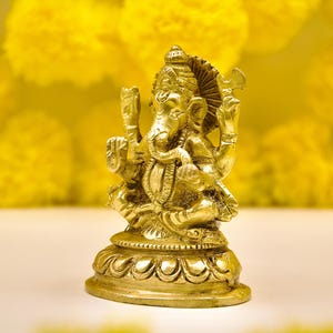 Puede incluir: Una estatua dorada de la deidad hind&uacute; Ganesha, sentado sobre una base decorativa. La estatua es muy detallada, con m&uacute;ltiples brazos y ornamentaci&oacute;n intrincada. El fondo presenta flores amarillas borrosas, creando un ambiente c&aacute;lido y acogedor.