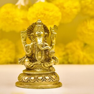 Puede incluir: Una estatua dorada de la deidad hind&uacute; Ganesha, sentada sobre una base decorativa. La estatua es muy detallada, con intrincadas tallas. El fondo presenta pompones amarillos borrosos, creando un ambiente c&aacute;lido. Es un art&iacute;culo religioso.