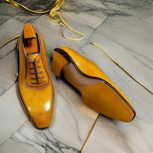 May include: A pair of mustard yellow leather oxford shoes with a polished finish, featuring dark brown laces, a rounded toe cap with decorative brogue detailing, and a low stacked heel. The shoes are displayed with one facing forward and one side-on.