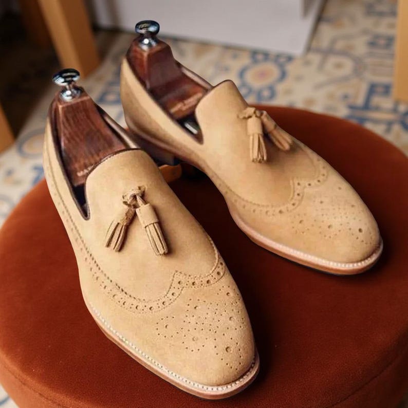 May include: A pair of tan suede loafers with decorative perforations and tassel accents. The shoes are displayed on a round, reddish-brown surface, showcasing their classic design and craftsmanship. The shoes have wooden shoe trees inside.