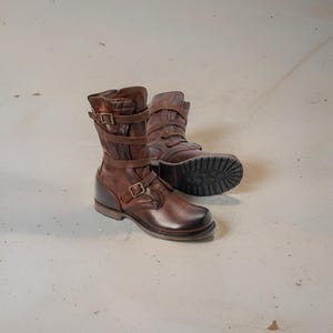 Puede incluir: Un par de botas de cuero marrón con múltiples correas y hebillas. Las botas tienen una apariencia robusta y desgastada, con costuras visibles y una suela texturizada. Las botas miden aproximadamente 25 cm de alto.