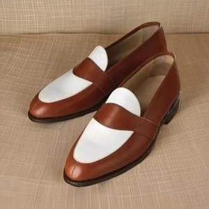 May include: A pair of two-tone loafers featuring a smooth brown leather body with a contrasting white leather saddle and vamp panel. These slip-on shoes have a rounded toe, a low heel, and a clean, polished finish across the entire exterior surface.