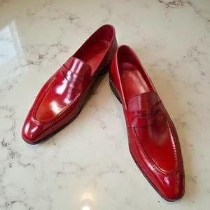 May include: A pair of glossy, red leather loafers. The shoes feature a classic penny loafer design with a rounded toe and a low heel. The leather has a polished finish, reflecting light. The shoes are displayed on a white marble surface.