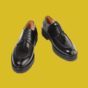 Op de afbeelding: Een paar zwarte leren derby-schoenen met een glanzende afwerking, voorzien van een moc-toe ontwerp, dunne zwarte veters en een dikke, grove zwarte profielzool. De binnenvoering van de schoenen is lichtbruin en zichtbaar bij de opening.