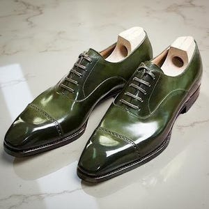 以下が含まれることがあります： 光沢のあるグリーンの革製ドレスシューズのペア。キャップトゥとレースアップクロージャーを備えたクラシックなオックスフォードデザインの靴です。木製のシューツリーが靴の中に入っています。