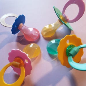 Könnte beinhalten: Eine Auswahl an bunten Babyschnullern in Rosa, Gelb, Blau und Grün. Jeder Schnuller hat einen abgerundeten Sauger und einen Ringgriff. Die Schnuller sind auf einer hellvioletten Oberfläche angeordnet.