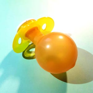 May include: A close-up of a baby pacifier. The pacifier has a yellow shield with two holes and a rounded orange nipple. The pacifier is made of plastic and is on a light blue surface.