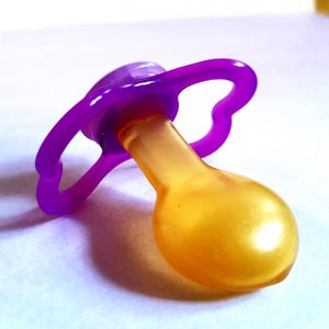 May include: A close-up of a baby pacifier. The pacifier has a purple ring and nipple, and an orange-yellow teat. The pacifier is made of plastic and is designed for infants.