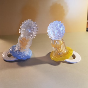May include: Two clear baby pacifiers. One is blue and the other is yellow. Each has a round, white handle. The top of each pacifier has a textured, spherical design.