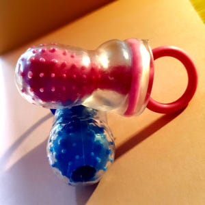 May include: A clear and pink baby fruit and vegetable feeder with a red handle. The feeder has a textured, pink, bulbous end and a clear, rounded body. A blue feeder is visible below.