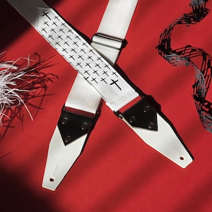 May include: White guitar strap with black cross pattern. The strap has black leather ends with metal rivets.
