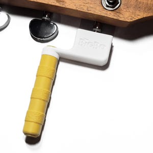 May include: A white plastic guitar string winder with a yellow textured handle and the embossed text BigBo, attached to the wooden headstock of a stringed instrument with visible chrome tuning pegs and a metal nut.