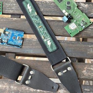 Op de afbeelding: Zwarte gitaarband met zilveren hardware en leren uiteinden. De band heeft een groene patch met een bliksemschicht en printplaatontwerp, en de tekst "BIGBO". Andere elektronische componenten zijn zichtbaar op de achtergrond.