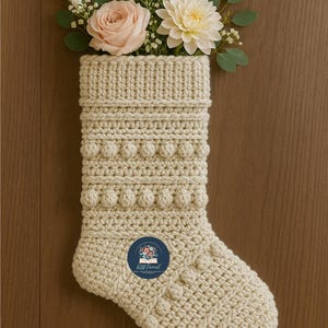 Peut inclure: Chaussette de Noël en crochet couleur crème, avec une boucle de suspension. La chaussette est ornée de points texturés et de boules. Un bouquet de fleurs, dont une rose rose et un dahlia blanc, est placé dans l'ouverture supérieure. Un autocollant avec du texte est sur la chaussette.