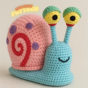 May include: A crocheted snail toy in shades of pink, blue, yellow, and green. The snail's shell is pink with red and purple details. The body is blue, and the eyes are yellow with green tops. The text "CROCHET PATTERN" is visible.