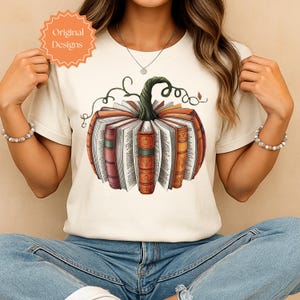 May include: Cream-colored t-shirt featuring a pumpkin design made of books. The pumpkin has a green stem and is surrounded by open books with orange, red, and brown spines. The text "Original Designs" is in a circular orange shape in the upper left.