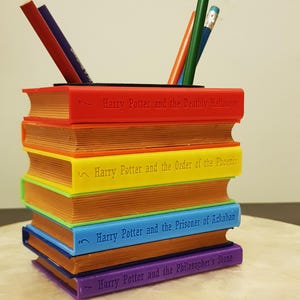 以下が含まれることがあります： ハリーポッターの本の積み重ねの形をしたカラフルなデスクオーガナイザー。本は赤、黄、青、紫で、背表紙にタイトルがエンボス加工されています。鉛筆とペンは一番上の本に置かれています。ユニークで機能的なデスクアクセサリー。
