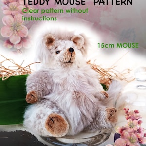 May include: A plush, light-colored teddy mouse, approximately 6 inches in height, sits on a clear glass surface. The image includes text that reads "TEDDY MOUSE PATTERN" and "15cm MOUSE". Decorative pink flowers and the text "PRINT IT ON A4" are also visible.