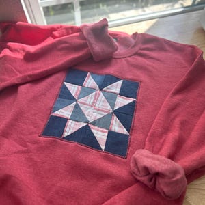 May include: A red long-sleeve sweatshirt featuring a central square quilt block design. The quilt pattern displays a star motif composed of dark blue denim and pink and white plaid fabric patches. A small rectangular label is stitched at the bottom.