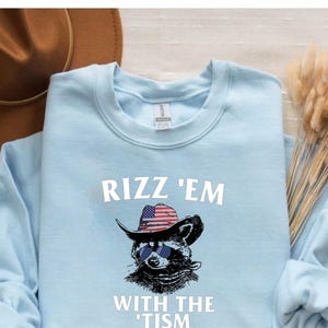 Può includere: Felpa blu chiaro con un procione che indossa un cappello da cowboy con una fascia con la bandiera americana e occhiali da sole. Il testo "Rizz 'Em With The 'Tism" è stampato sulla felpa.