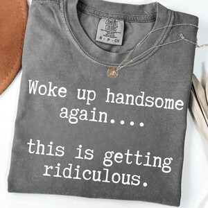 May include: A charcoal grey short-sleeve t-shirt with a crew neckline. The shirt features white printed text that reads, "Woke up handsome again.... this is getting ridiculous." A small gold-toned pendant necklace rests on the collar area.