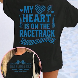 Puede incluir: Camiseta negra con el texto azul "MY HEART IS ON THE RACETRACK". El diseño incluye un corazón a cuadros, un rayo y banderas de carreras. Una imagen más pequeña muestra el texto "RACE AUNT ERA".