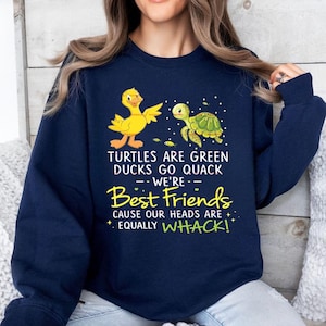 以下が含まれることがあります： 黄色いアヒルと緑のカメのグラフィックが特徴のネイビーブルーのスウェットシャツ。「TURTLES ARE GREEN DUCKS GO QUACK -- WE'RE -- Best Friends CAUSE OUR HEADS ARE + EQUALLY WHACK!」と書かれたテキスト。