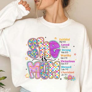 Puede incluir: Sudadera blanca con las palabras "She is Mom" en letras coloridas y con burbujas. La palabra "She" tiene un estampado floral, y "Mom" tiene un patrón ondulado. El texto adicional incluye "Faithful", "Loved", "Strong", "Worthy", "Victorious", "Blessed" y "Cherished".