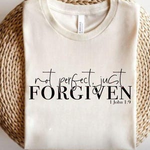 May include: Cream-colored t-shirt with the words "not perfect, just FORGIVEN" in black script and block letters. The text "I John 1:9" is below the main text. The shirt is displayed on a woven, circular surface.